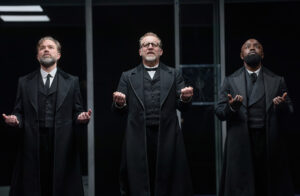Production photo from the National Theatre production of The Lehman Brothers showing three actors holding their hands out 
