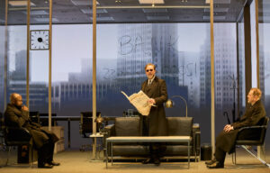 Production photo from The Lehman Trilogy February 2023 showing an actor standing reading a newspaper with two other actors sitting each side of him