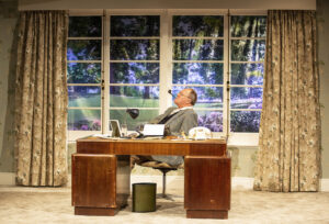 Production photo from the play Wodehouse In Wonderland February 2023 shows the actor Robert Daws istting at a desk with a typewriter next a window with a sunny view