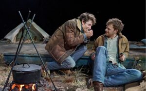 Actors Mike Faist plays a harmonica while he and Lucas Hedges crouch round a camp fire in the stage production of Brokeback Mountain at sohoplace theatre in London in June 2023