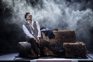 Danny Mac seated on a bale of straw with smoke around him while playing John Wilkes Booth in the Chichetser Festival Theatre production of Stephen Sondheim's Assassins in June 2023
