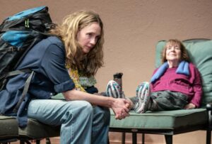 Two women sit next to one another, one is wearing a rucksack, in a stage production of infinite Life by Annie Baker
