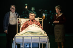 A scene from Nye at the National Theatre London in which actor Michael Sheen is lying on a hospital bed with actors Roger Evans and Sharon Small standing either side of him