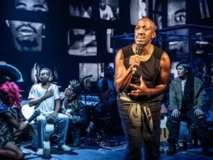 Actor Marc Brenner stands upstage with a microphone in front musicians and bideo screens in scene from the Young Vic production of Passing Strange