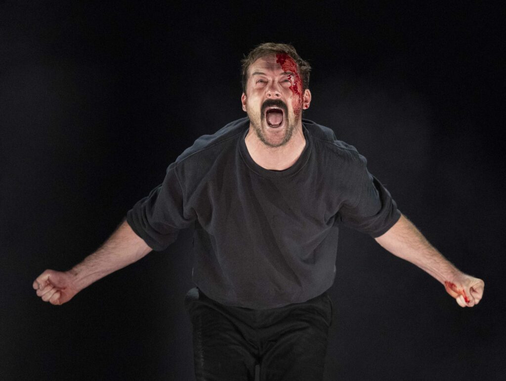An bloodied male actor stands with his arms open in a scene from the stage prodcution of Boys From The Blackstuff at the Garrick Theatre Kondon