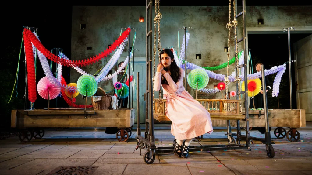 A young female actor sits on swing surrounded by balloons and hanging paper decorations in a scne from The Secret Garden at the Open Air Theatre in Regent's Park London