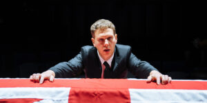 A man in suit leans across a coffin draped in an England flag in a scene from Death of England: Michael at sohoplace 