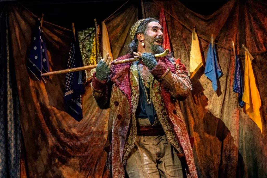 A smiling bearded actor in a colourful outfit holds his walking stick like a flute